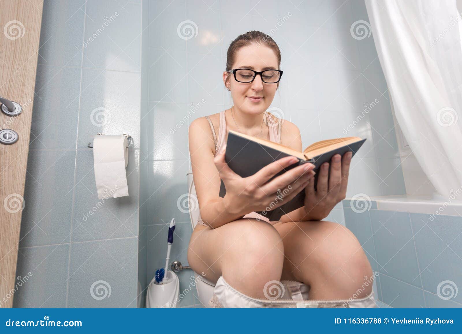 Stående av den unga nerdy kvinnan med glasögon som sitter på toalett och läseboken. Ung nerdy kvinna med glasögon som sitter på toalett och läseboken