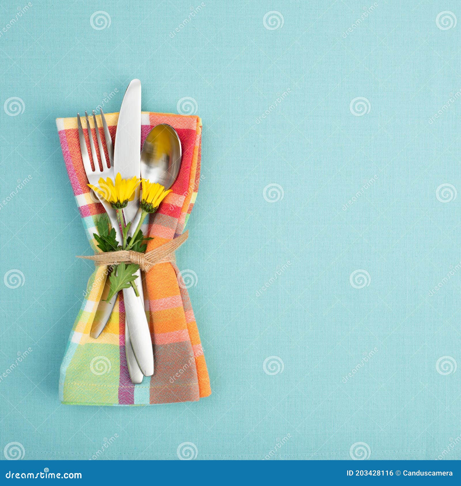summer or spring table setting with teal tablecloth, silverware, and multicolor cloth napkin with yellow daisy flowers.  backgroun