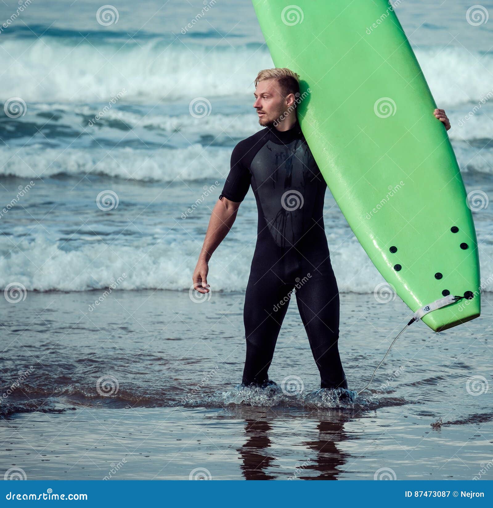 Surfer Wearing Wetsuit Standing on the Beach with a Surfboard Stock