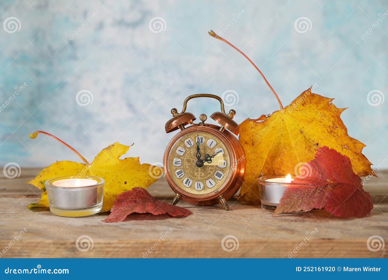 switching from summer to winter time, vintage alarm clock showing the set back of one hour, autumn leaves and candles on rustic