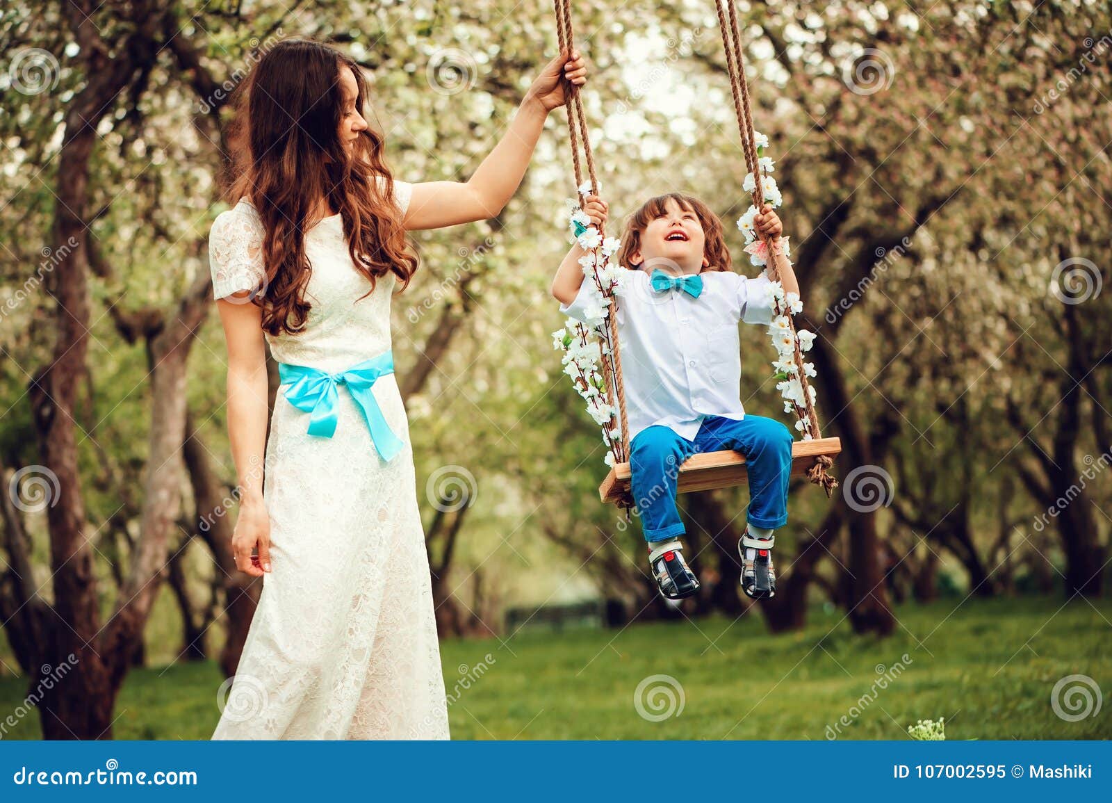 Szczęśliwy dressy matki, berbecia dziecka syn ma zabawę na huśtawce w parku i, będący ubranym łęku krawat i długą koronkową suknię dla urodziny lub matka dnia