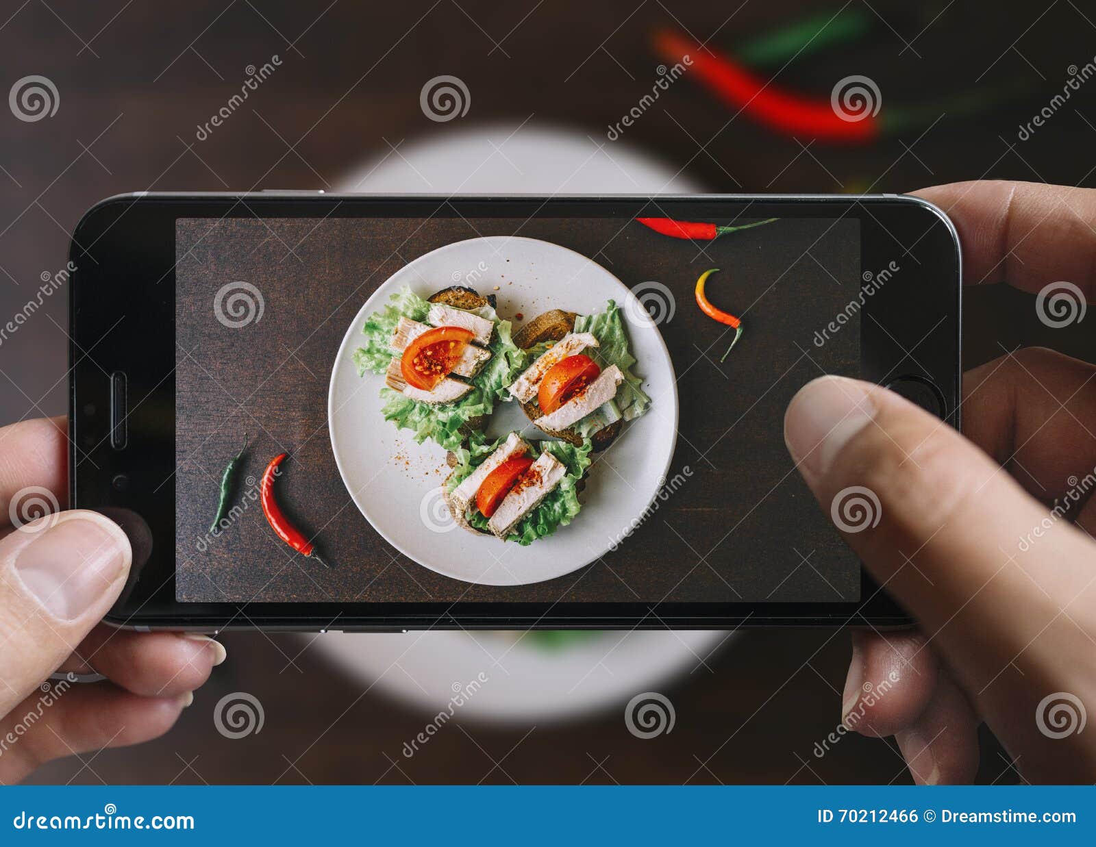 Ta bilden av skinksmörgåsen med mobiltelefonen Telefon i manliga händer På plattan finns det 3 skinksmörgåsar med tomaten och grönsallat Plattan är på en svart åldras trätabell tappning för stil för illustrationlilja röd