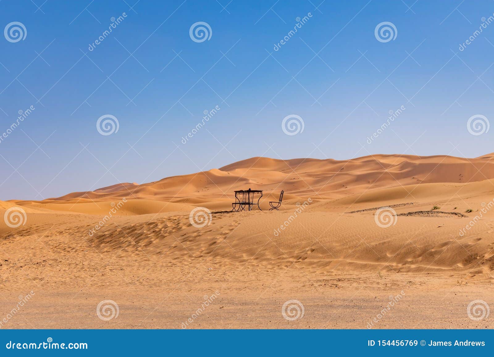 Tabela e cadeira na distância em uma duna de areia em Sahara Desert. Uma tabela simples e uma cadeira em uma duna de areia em Sahara Desert em Marrocos com um céu azul