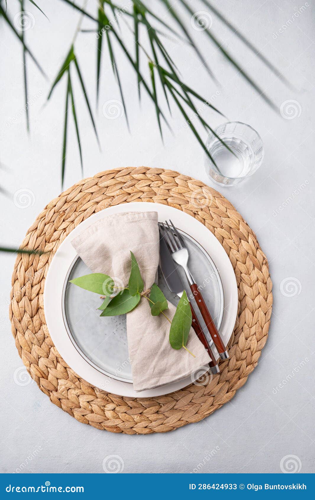 Table Setting with a Blue Plate and a Glass on a Straw Napkin with Plant Branches on a Light