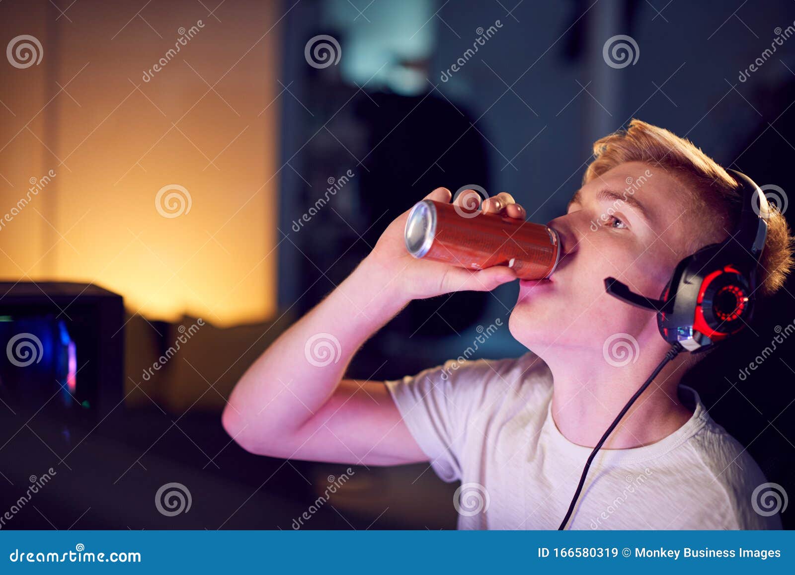 teenage boy drinking caffeine energy drink gaming at home using dual computer screens at night