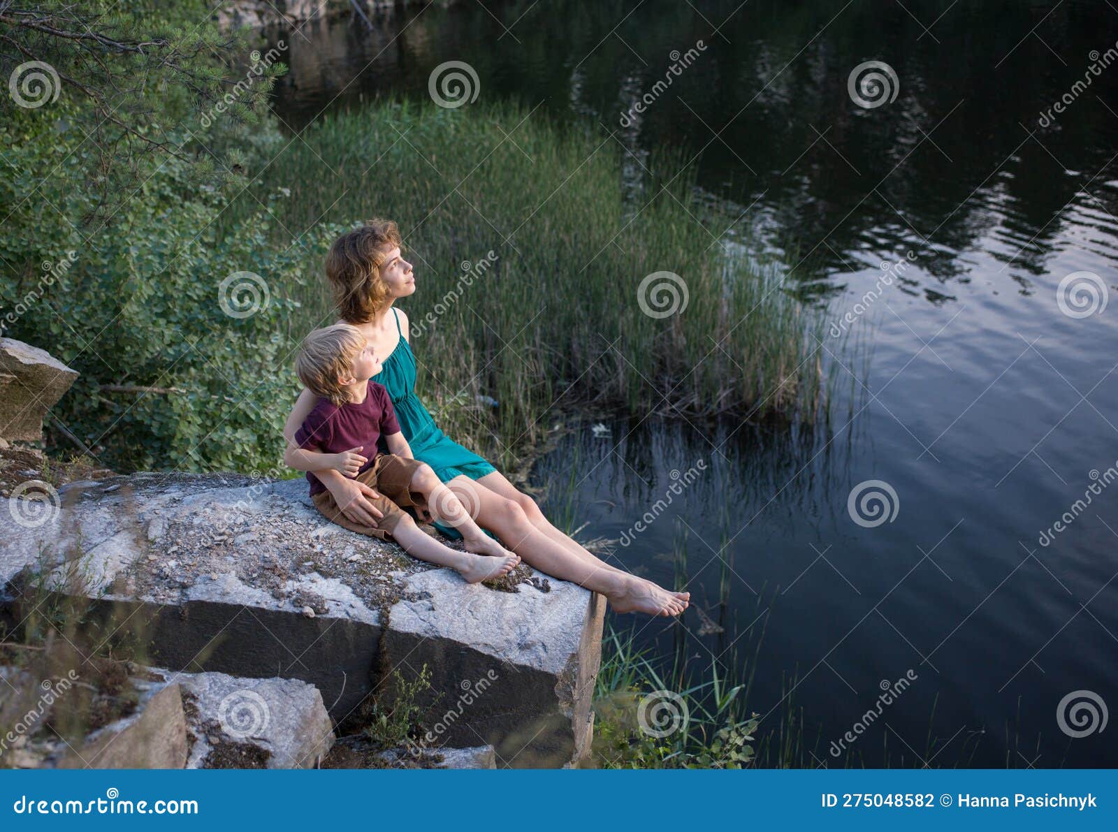 teenage girl and a boy, brother and sister, are happily sitting together on rock near lake and fantasize with interes