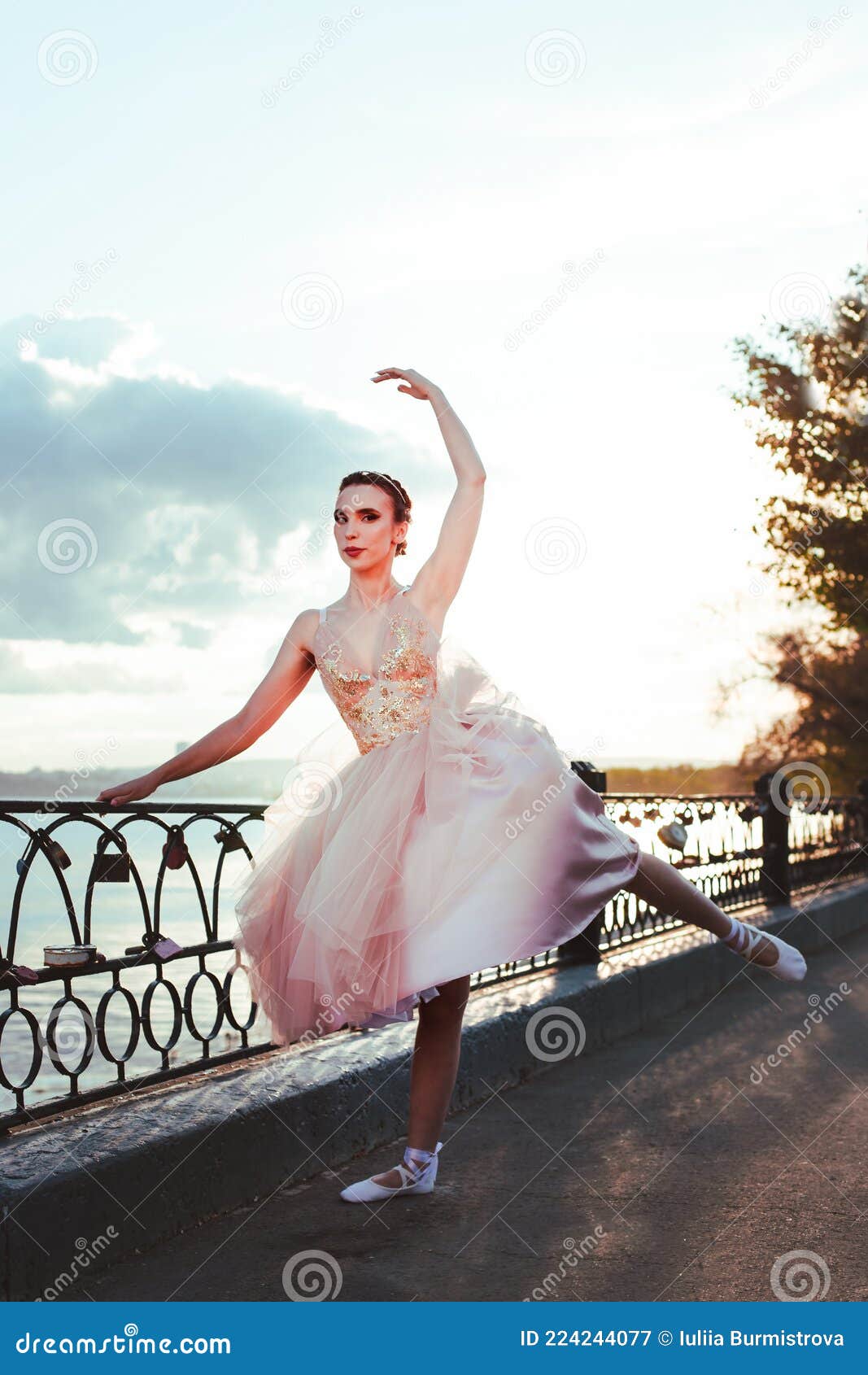 a thin ballerina in a pink silk corset dress gracefully raises her pointe-toed leg back on the river embankment in a