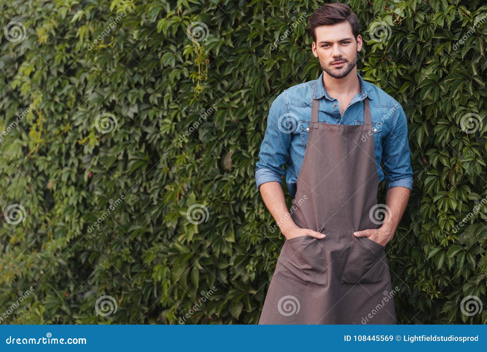 thoughtful gardener in apron looking at camera while standing in garden