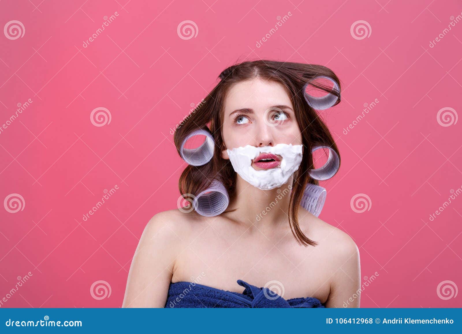 A thoughtful girl, with curlers on hair and with shaving foam on face, looks up with her mouth open. A thoughtful young girl, with large curlers on her hair and with shaving foam on her face, looks up with her mouth open. On a pink background.