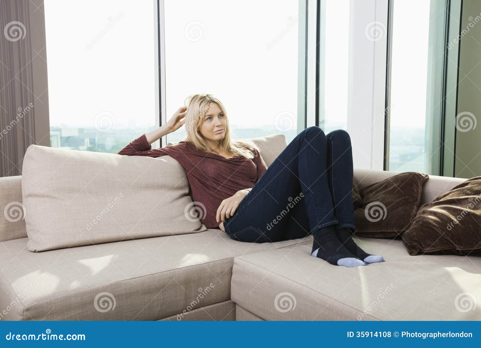 thoughtful young woman sitting on sofa against window at home
