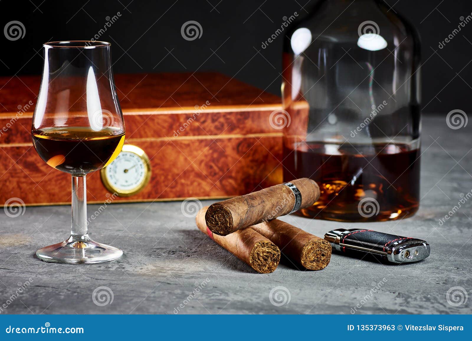 three cuban cigars on a stone table with a lighter and a wooden humidor with a glass and a bottle of whiskey