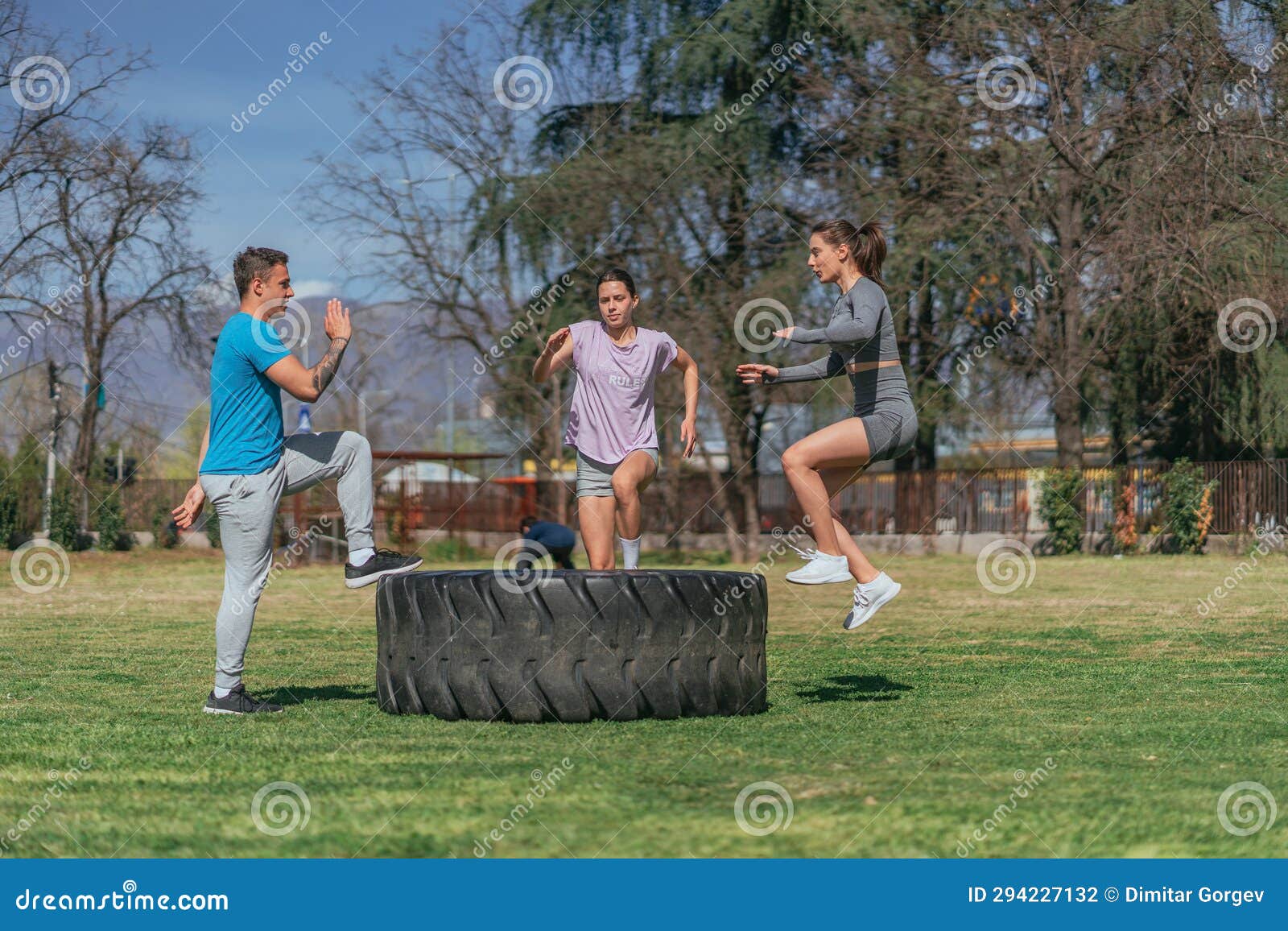 three sports friends doing jumps on a giant truck tire. they are exercising in the park, on a sunny day