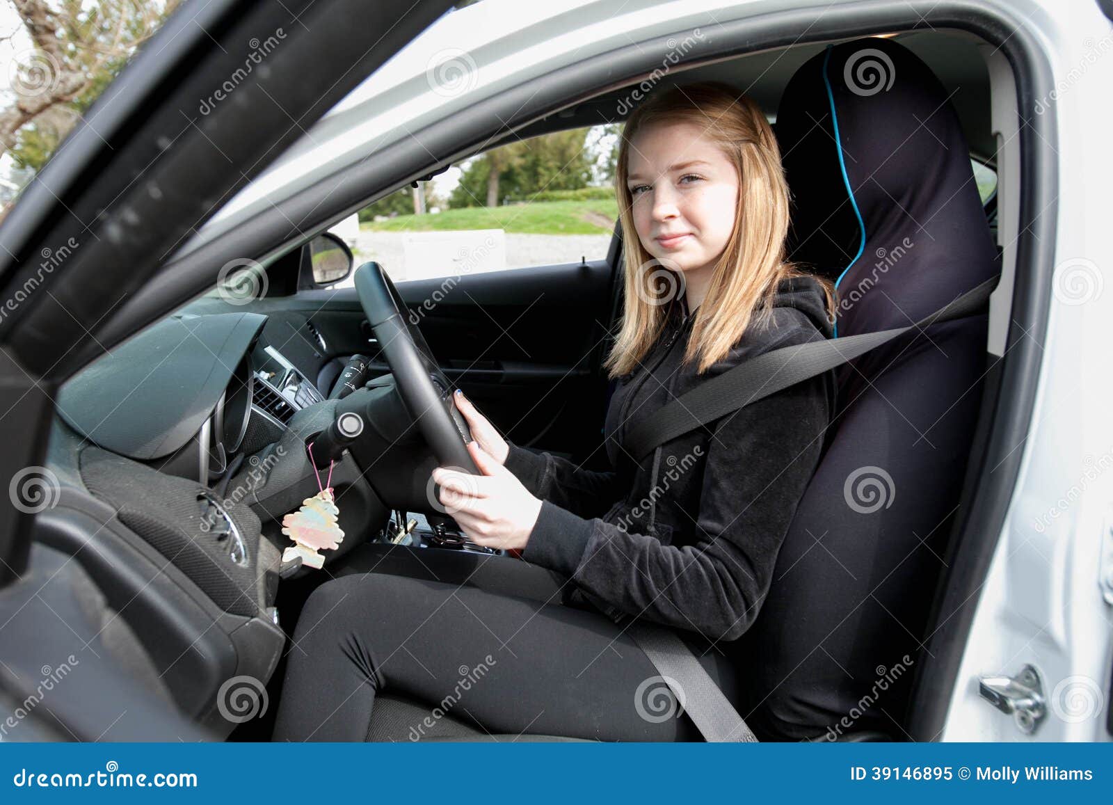Tienerbestuurder in auto. Tienerzitting in de bestuurderszitplaats van een auto met haar die handen op het stuurwiel, vanuit het perspectief van de open autodeur wordt genomen.