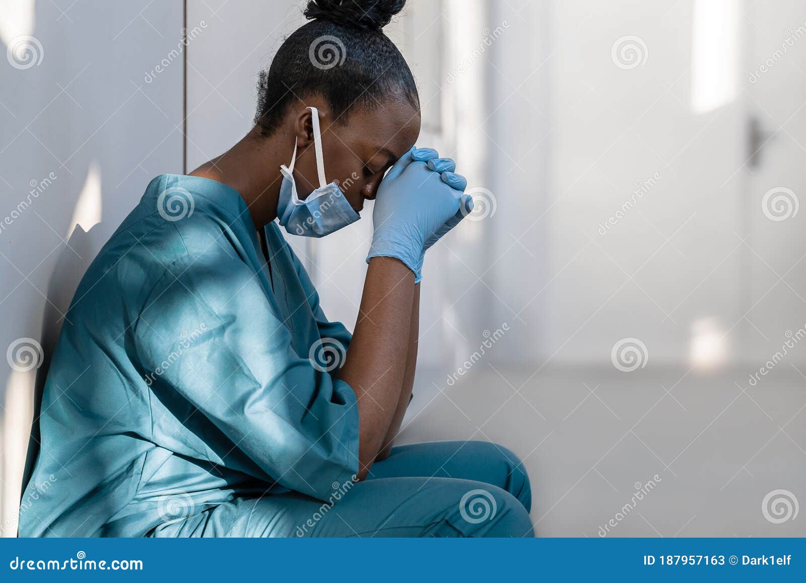 tired depressed female african nurse wearing face mask sits on hospital floor.