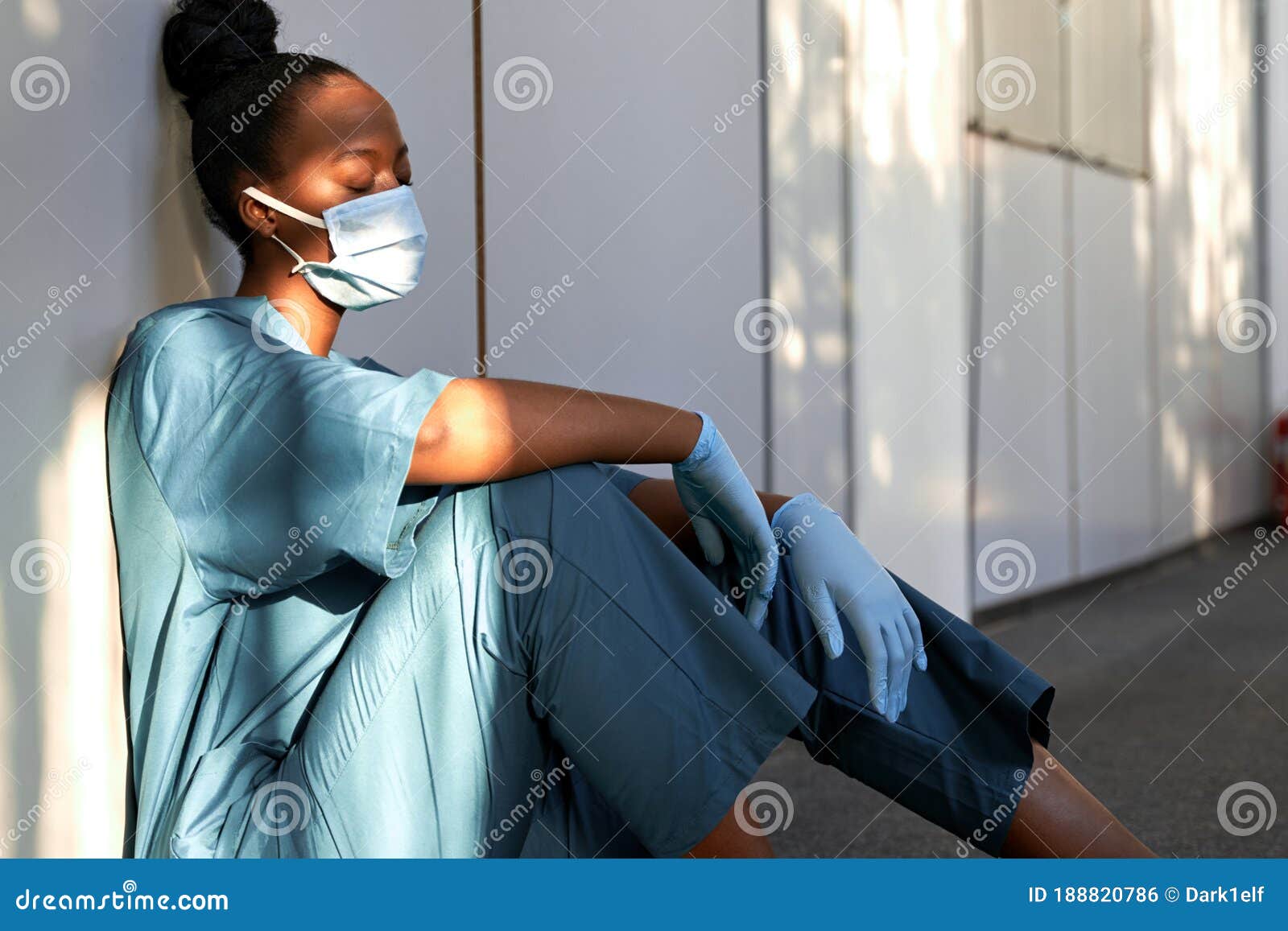 tired exhausted woman african nurse wear face mask gloves sit on hospital floor.