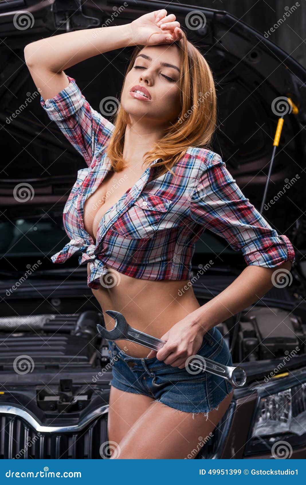 Tired after hard working day. Beautiful young woman with beautiful cleavage holding big wrench and keeping eyes closed while standing near the car in auto repair shop