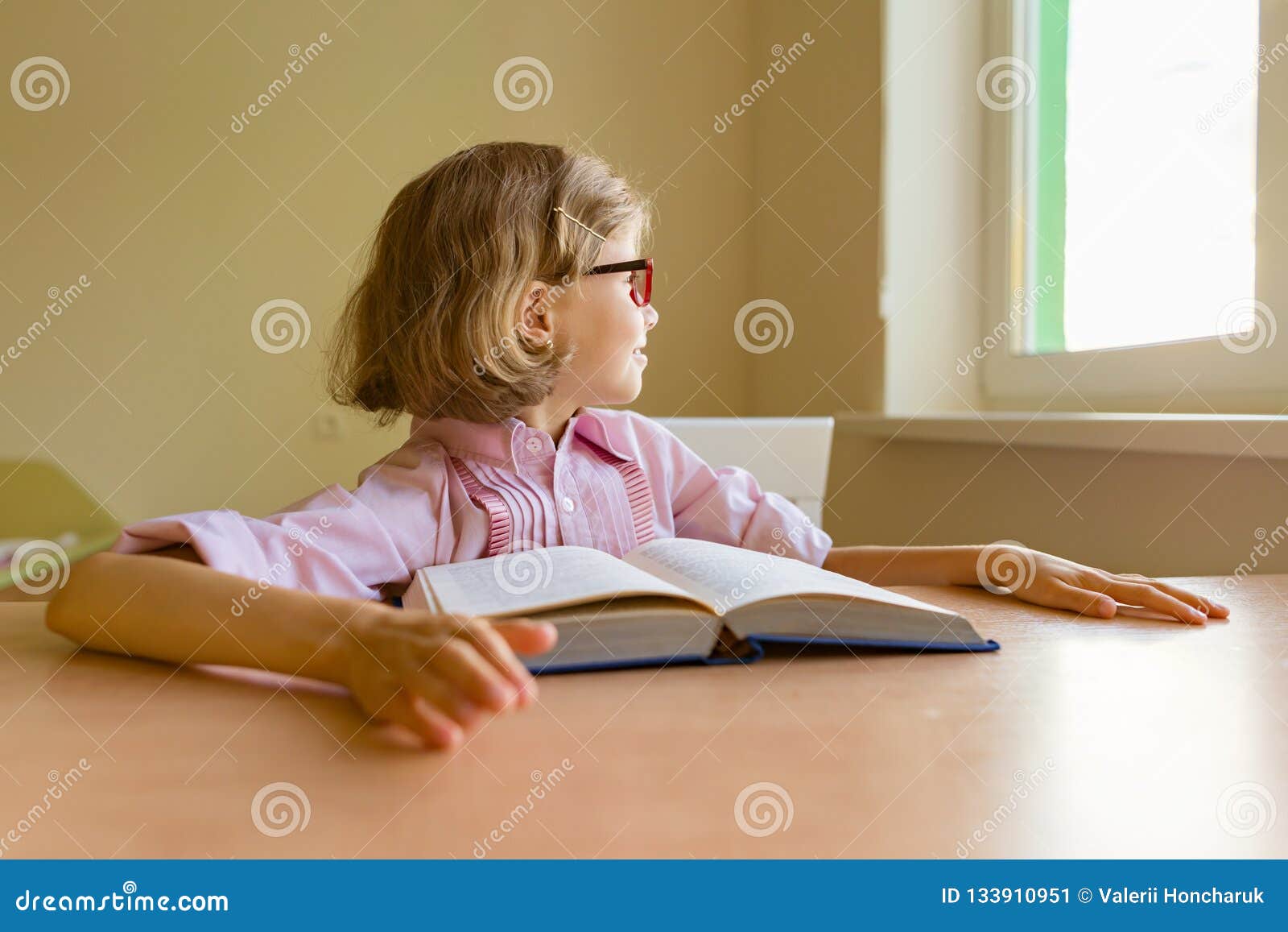 Tired little girl student looks out the window while sitting at her desk with a big book. School, education, knowledge and children.