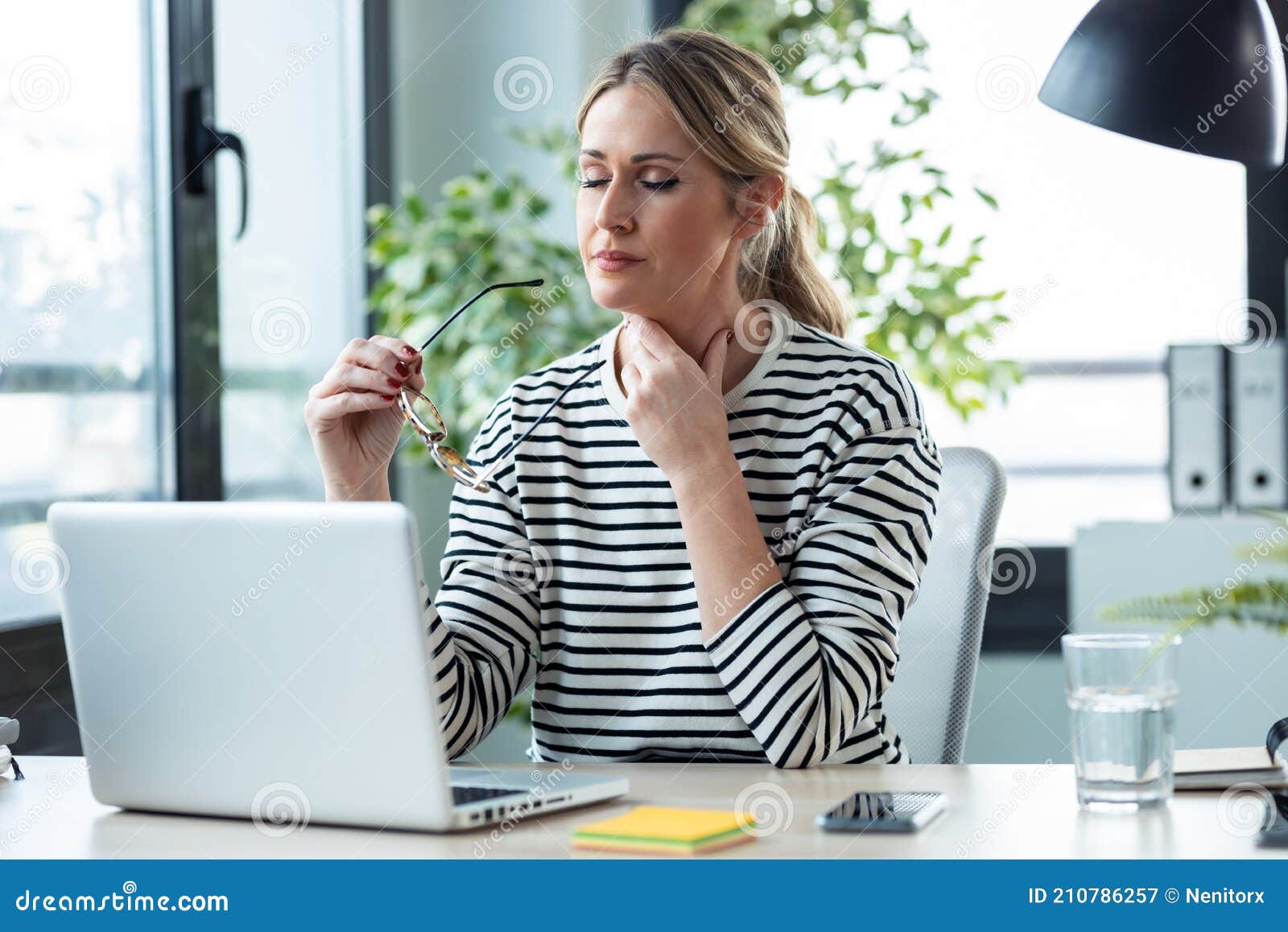 tired mature business woman with a sore throat looking uncomfortable while working with laptop on a desk in the office at home