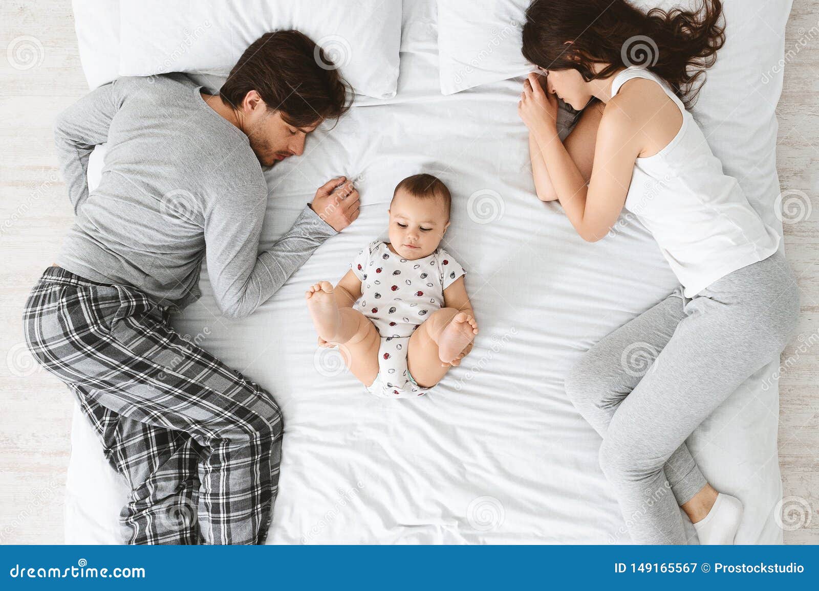 Tired Parents Sleeping on Bed Sides, Baby Playing in the Middle Stock