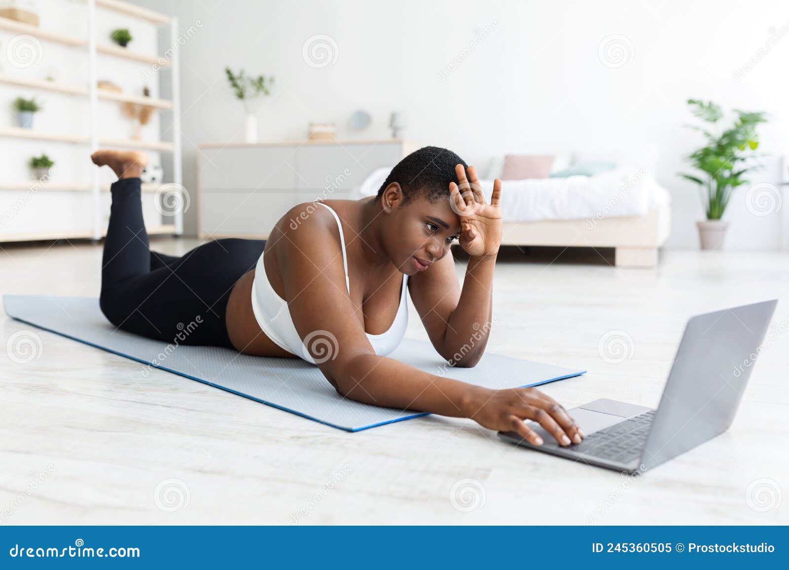 Tired Plus Size Black Woman Lying on Yoga Mat Near Laptop, Wiping