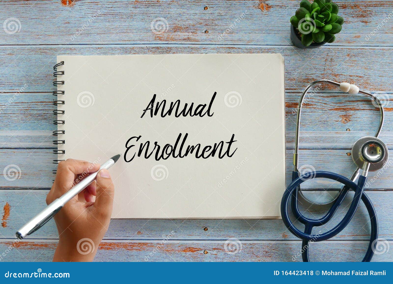 top view of plant,stethoscope, and hand writing annual enrollment on notebook on wooden background
