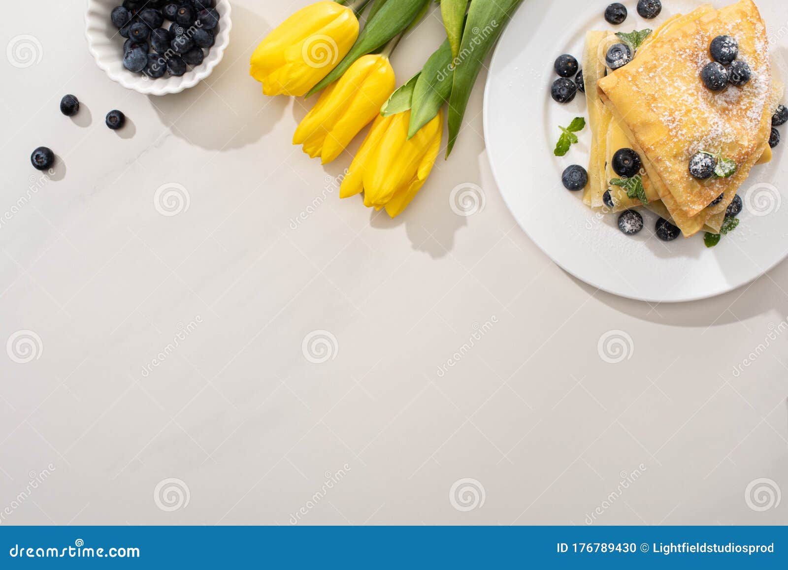 Top view of tasty crepes near bowl of blueberries and yellow tulips on grey background