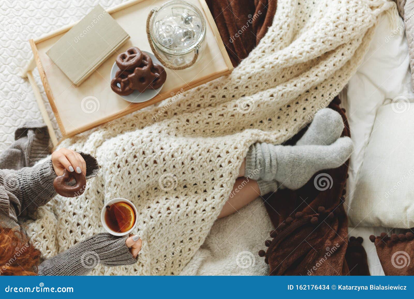 top view of young girl covered with cozy white blanket siting on bed drinking tea and eating
