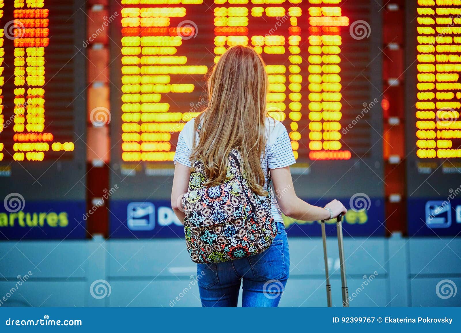 tourist girl with backpack and carry on luggage in international airport, near flight information board