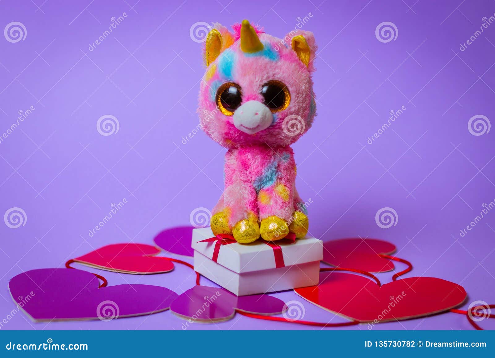 Toy Pink Unicorn Sits On A White Gift Box With A Red Ribbon On Paper Hearts On A Purple