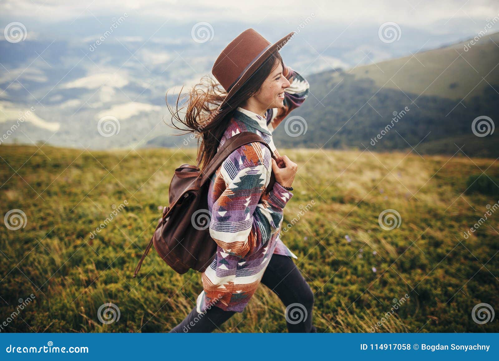 travel and wanderlust concept. stylish traveler hipster girl holding hat, with backpack and windy hair, walking in mountains in c