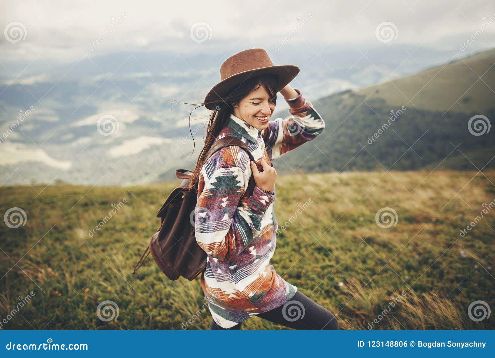 travel and wanderlust concept. stylish traveler hipster girl holding hat, with backpack and windy hair, walking in mountains in c