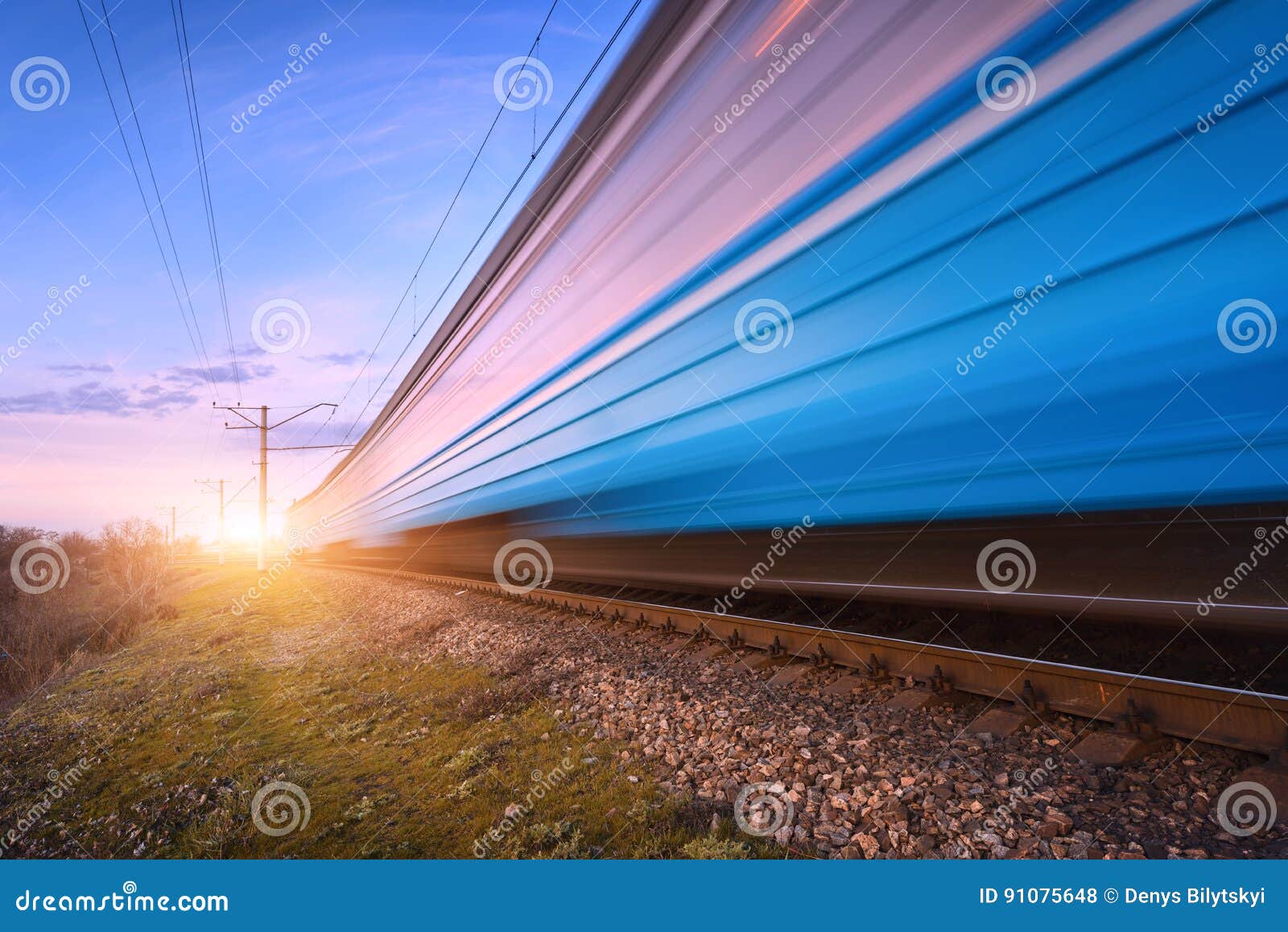 Treno passeggeri blu ad alta velocità nel moto sulla ferrovia al tramonto Treno pendolare moderno vago Stazione ferroviaria e cielo variopinto Viaggio della ferrovia, turismo ferroviario Paesaggio industriale rurale nel crepuscolo