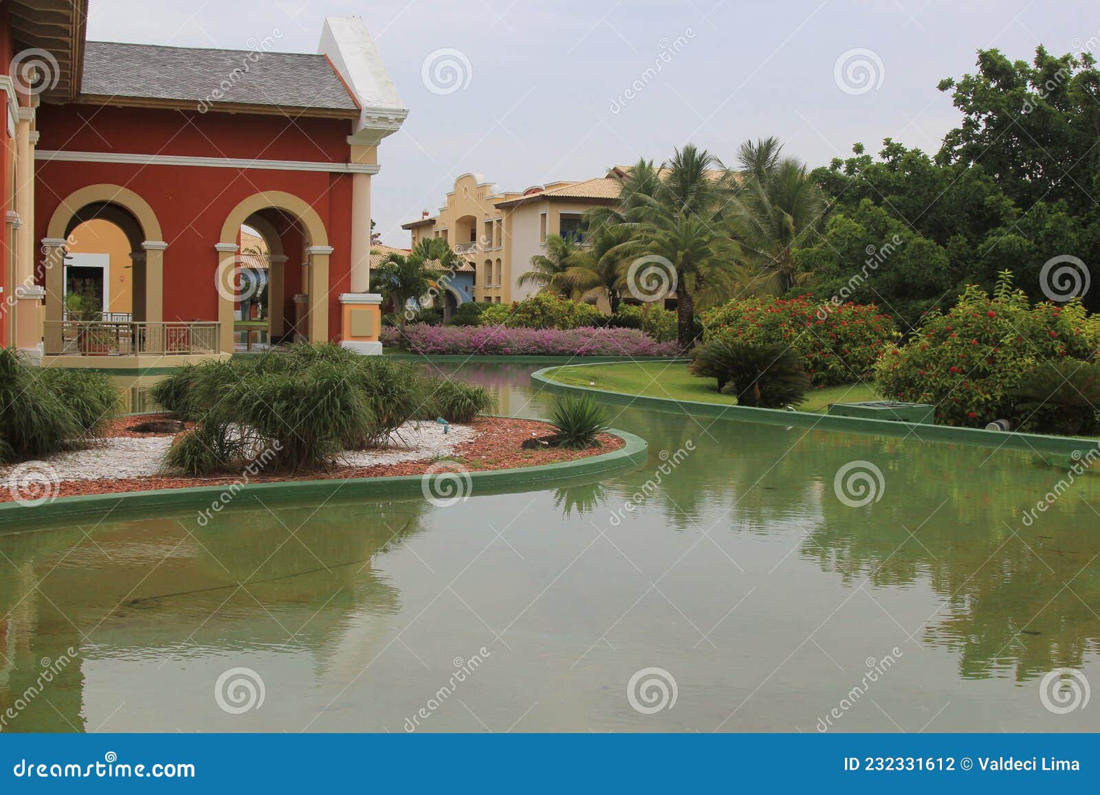 tropical garden with green grass, colorful houses in the background, a small lake with calm water and reflections abrir no google