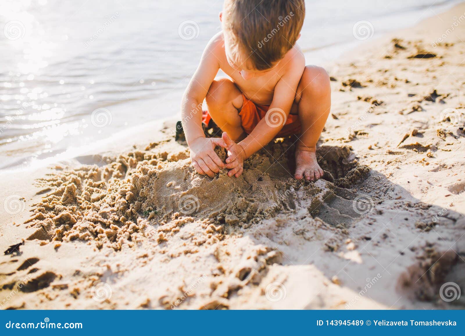 A little boy builds figures from the sand on the shore of the pond at sunset of the day, hands dig up the sand in a crisp plan