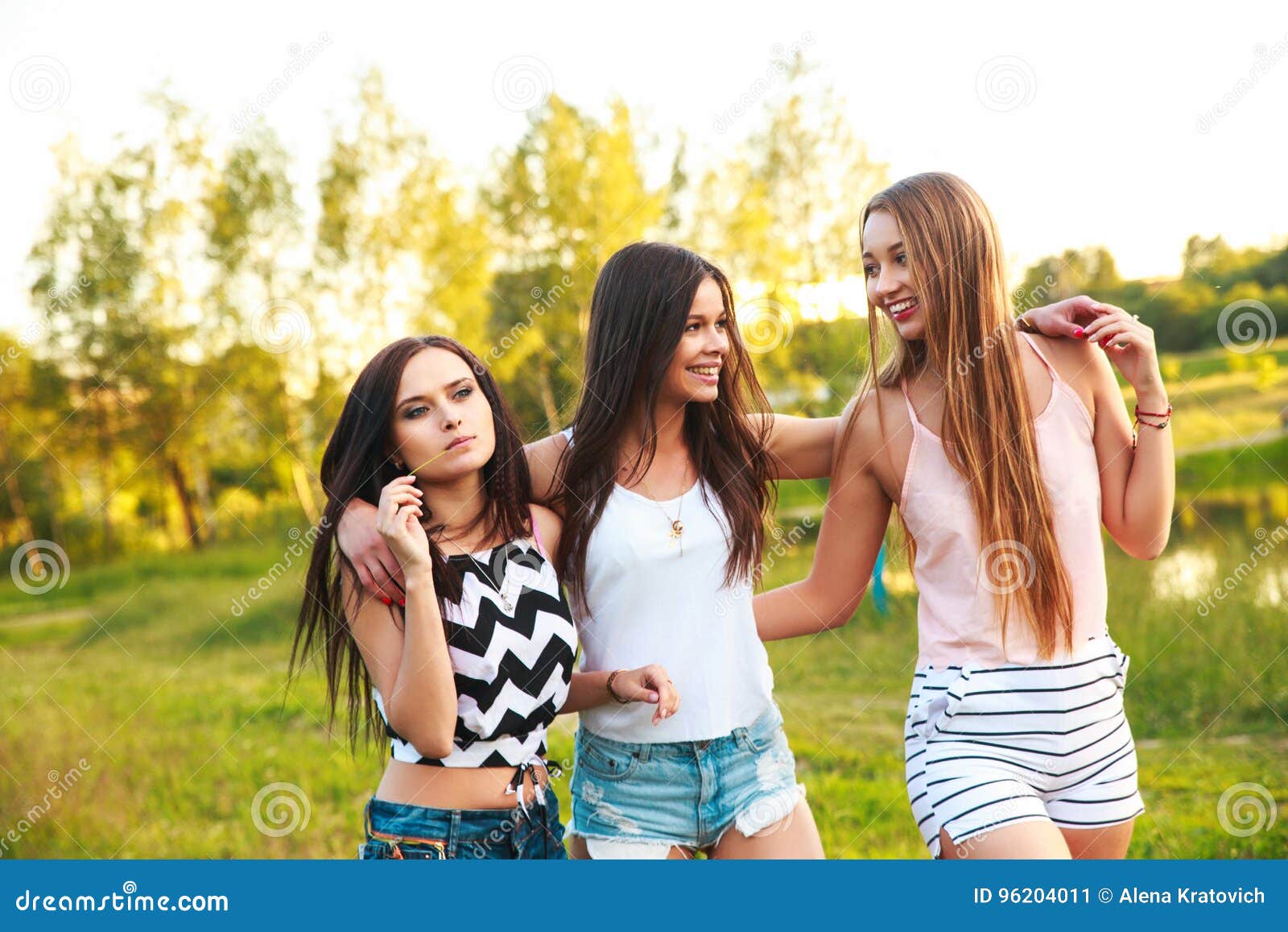 Três meninas bonitas que andam e que riem no por do sol no parque Conceito da amizade. Três mulheres que abraçam-se fora e que riem no por do sol summertime