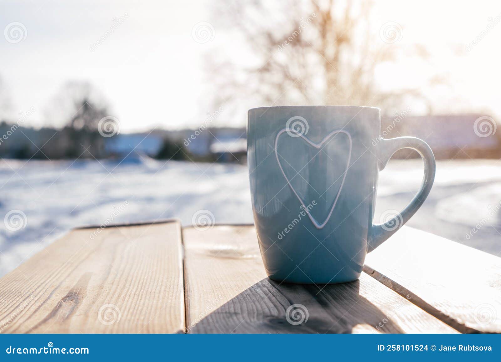 turquoise blue cup with heart rising steam on wooden table against backdrop of trees and houses with snow on winter day