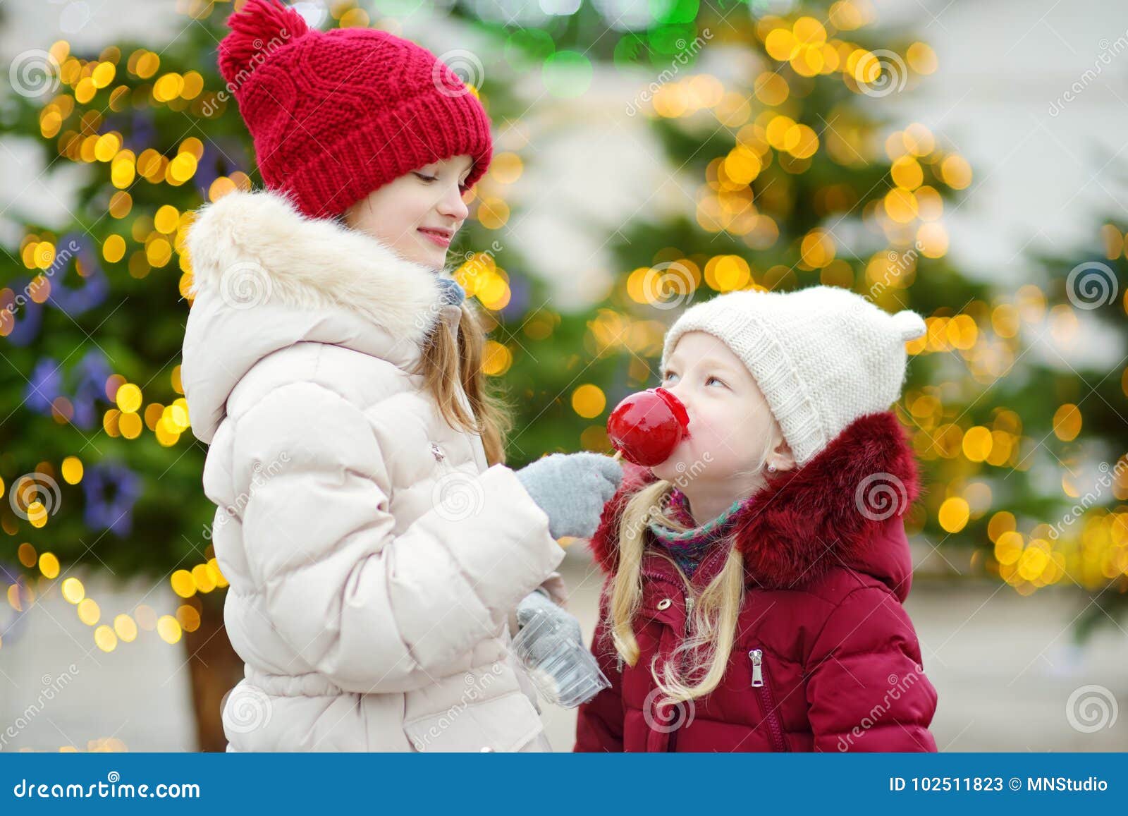 Two adorable little sisters eating red apples covered with sugar icing on traditional Christmas market Children enjoying sweets can s and gingerbread on