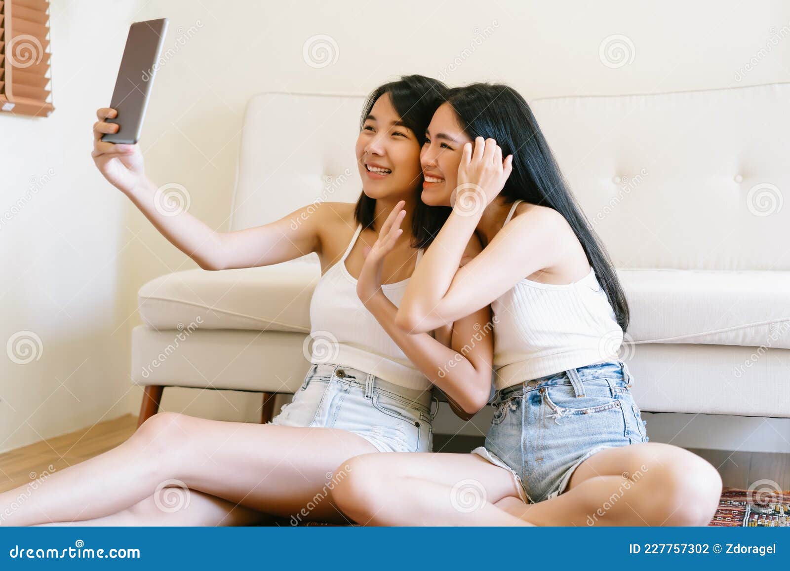 Two Beautiful Asian Girls or Lesbian Couple Setting on the Floor and Waving Hands Making Distance Video Call Looking at Digital Stock Photo
