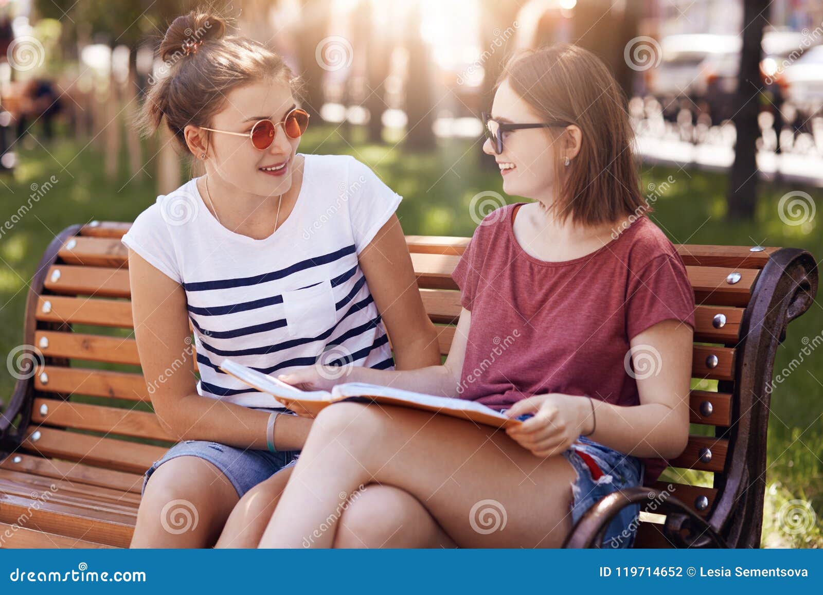two beautiful females students wears summer shades, rest on bench in park, have positive expressions, cramm for exam, discuss futu