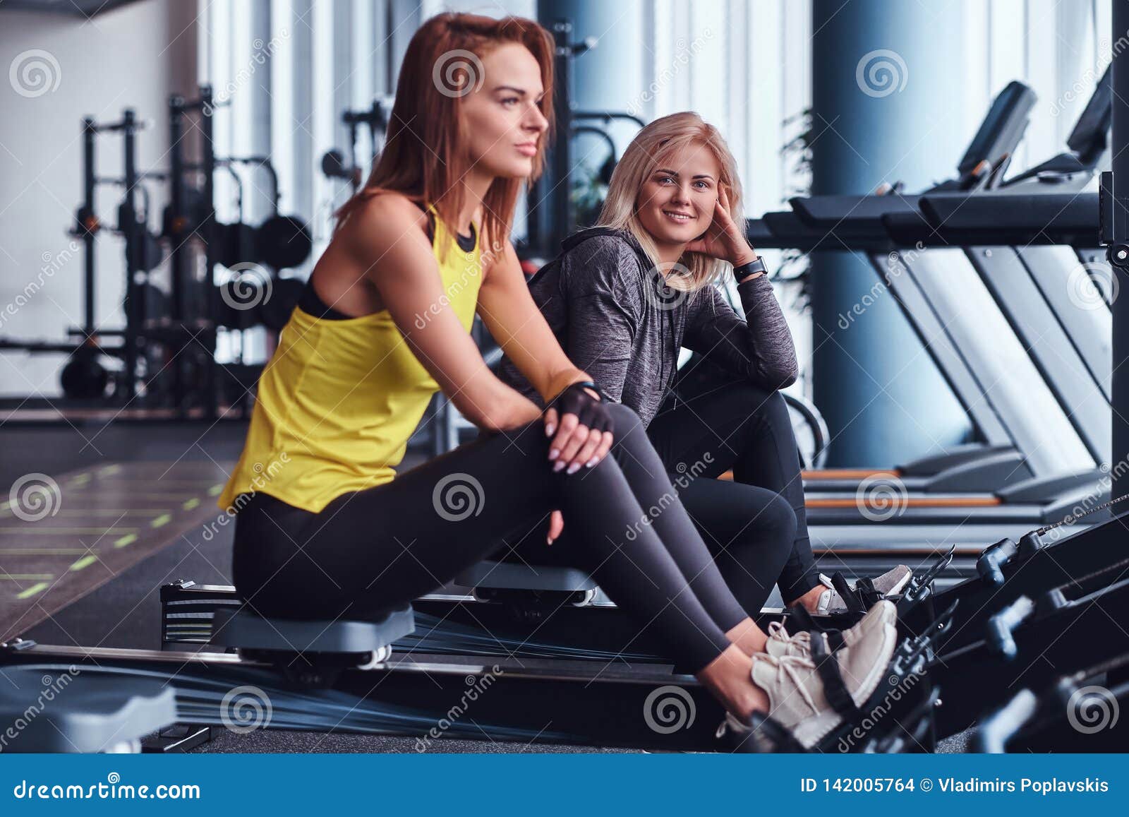 Two Beautiful Young Women Sitting On Rowing Machines At The Gym. Stock ...