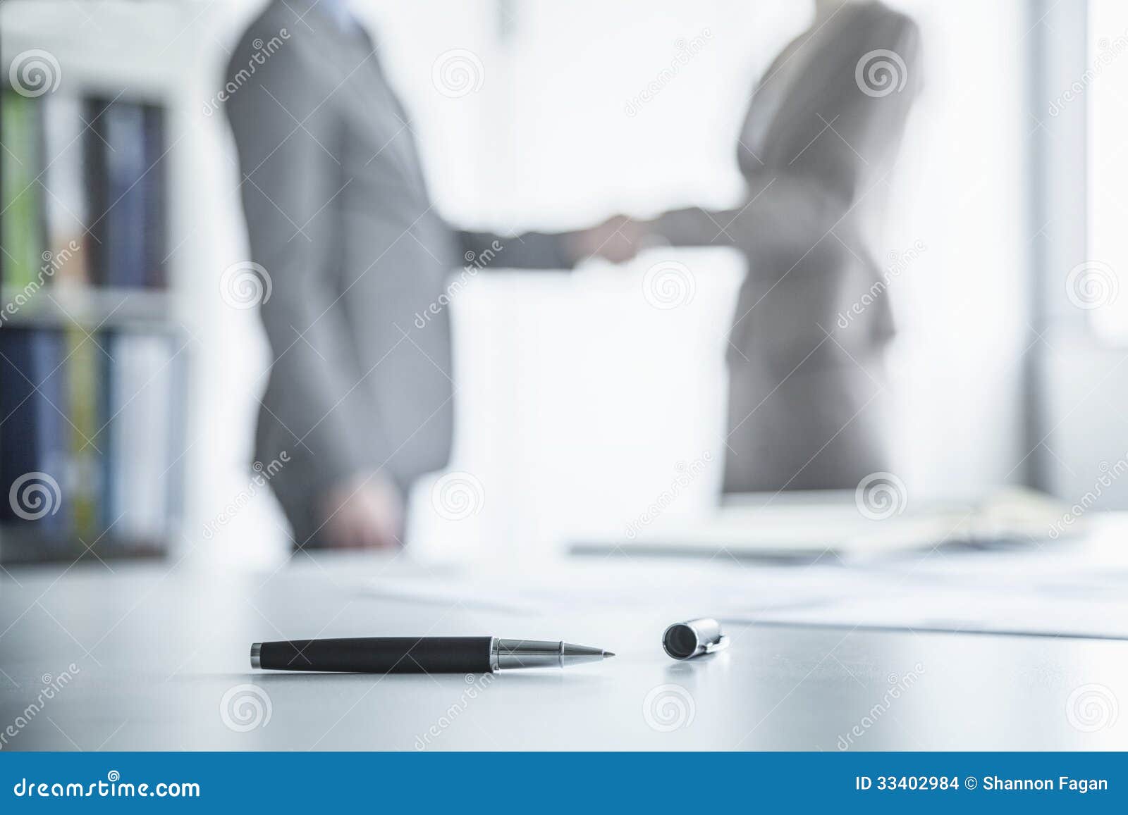 two business people shaking hands in the background, pen lying on the table in the foreground