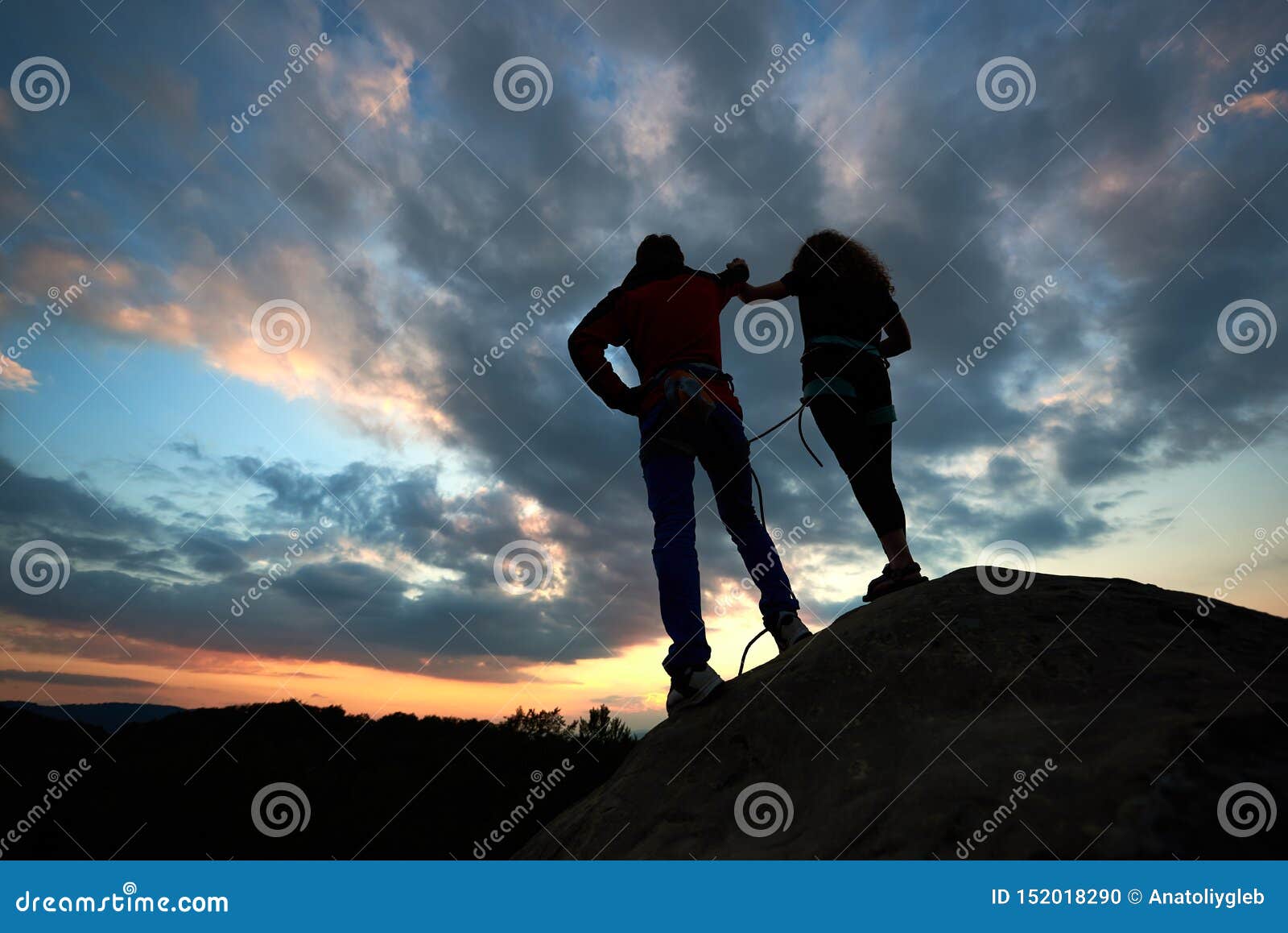 silhouettes of man and woman look at sunset on the top of rock. hikers couple on dramatic sky at sunset. back view.