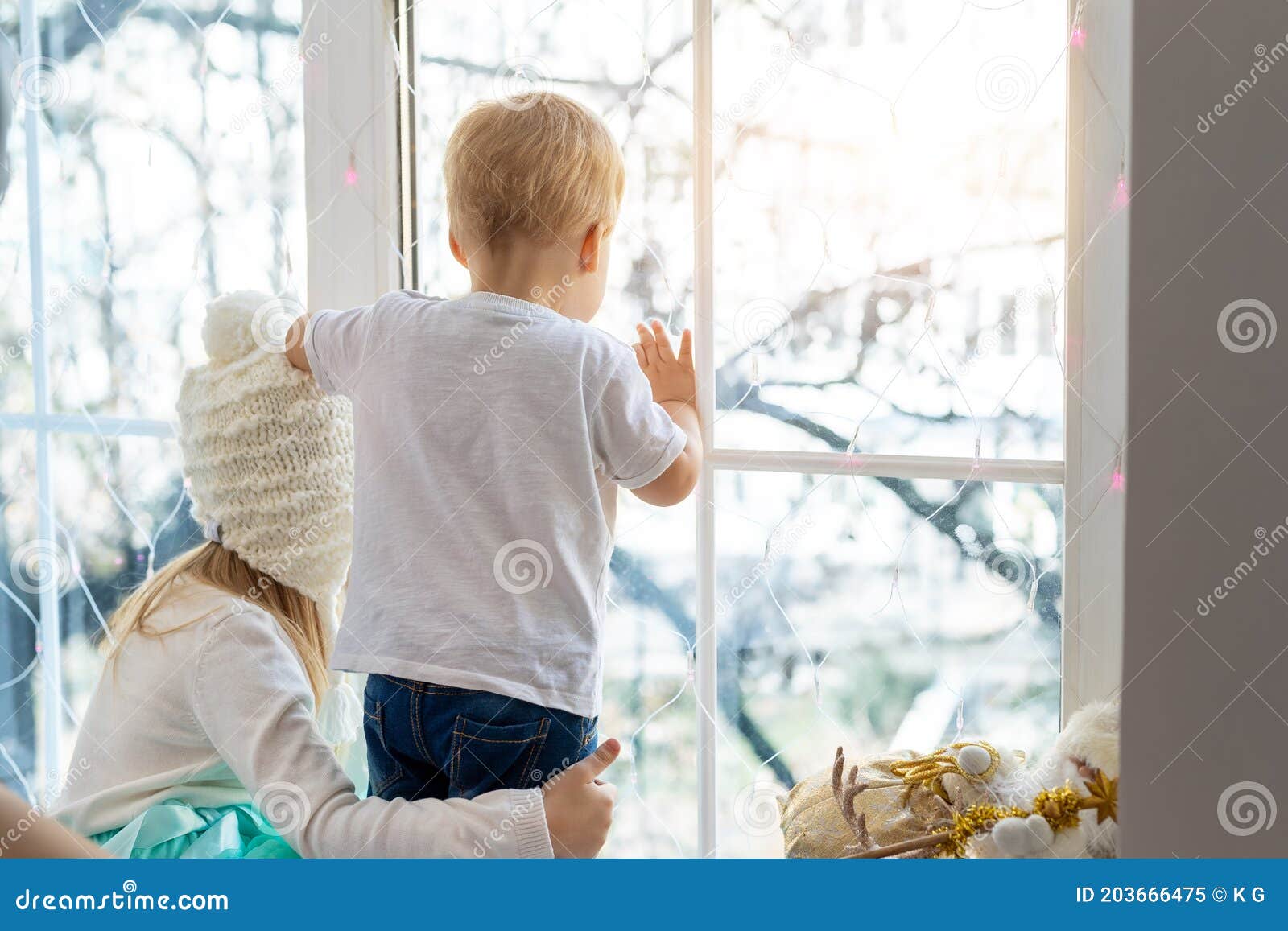 two cute adorable little blond cauasian children siblings stay near window and looking outside waiting for snow, wonders