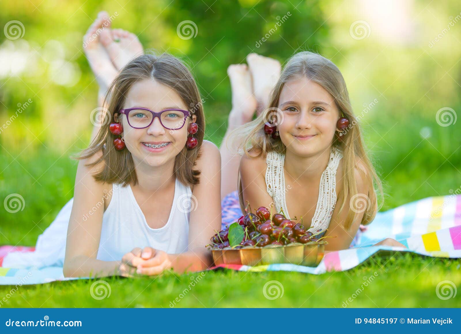 Two Cute Sisters Or Friends In A Picnic Garden Lie On A Deck And Eat Freshly Picked Cherries