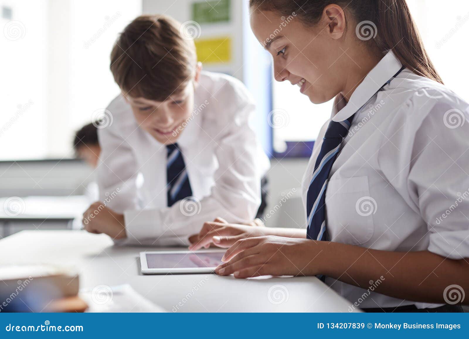two high school students wearing uniform working together at desk using digital tablet
