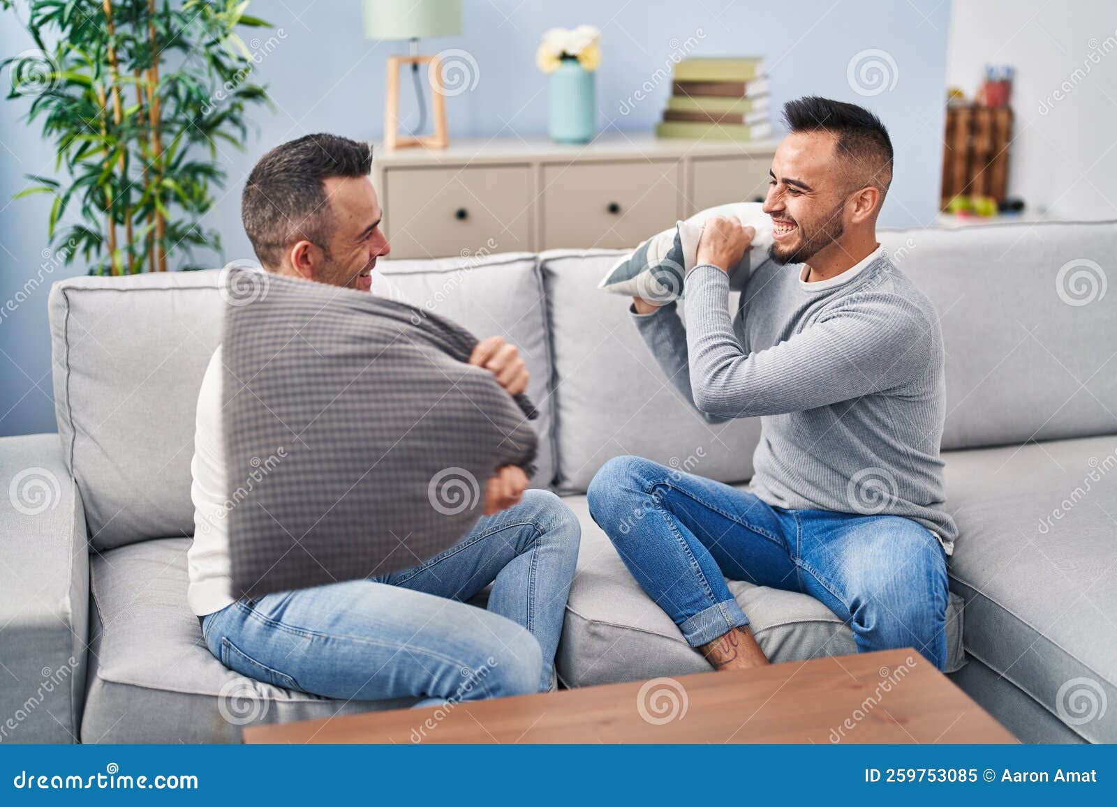 Two Men Couple Fighting with Cushion Sitting on Sofa at Home Stock