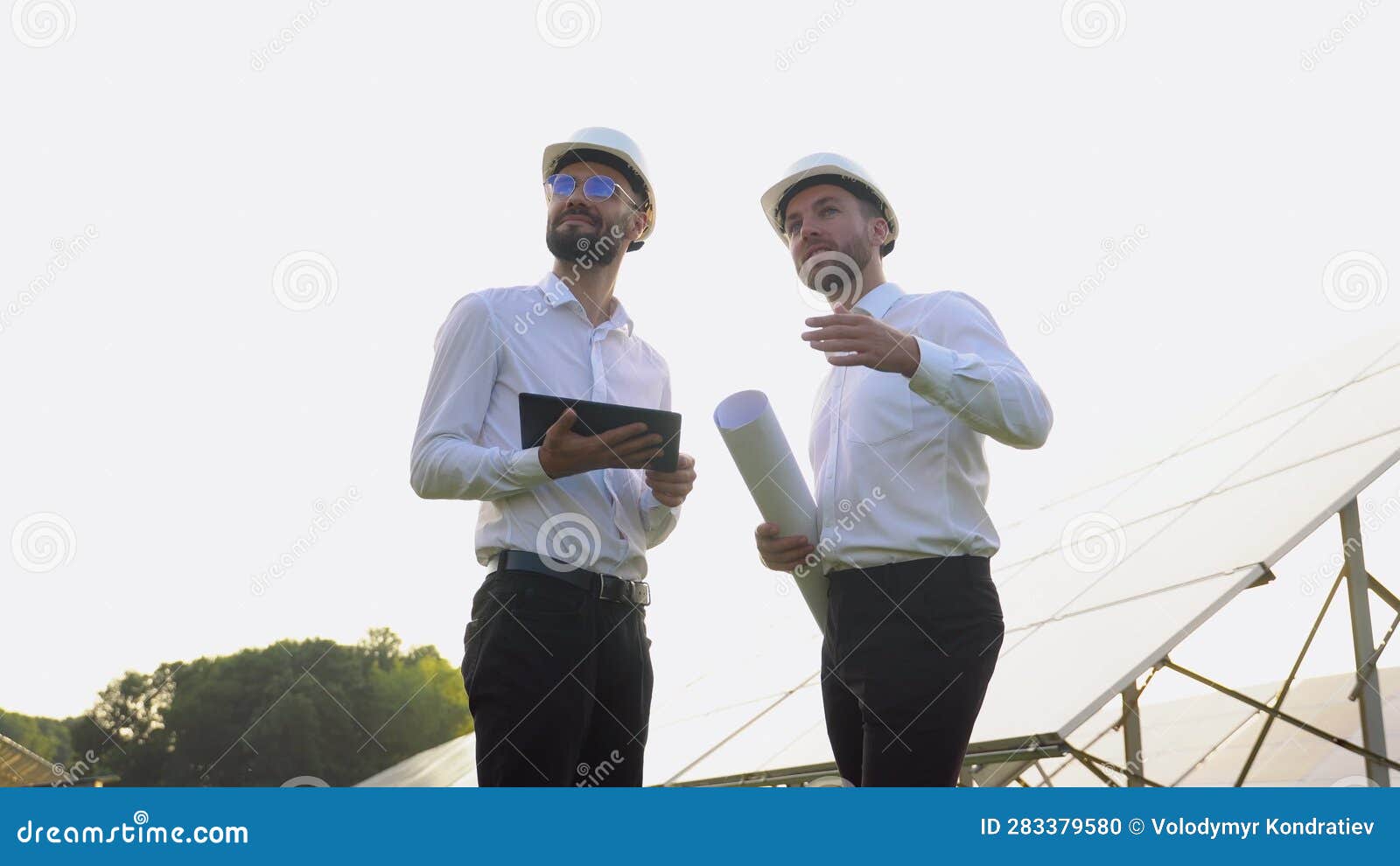 Two Men in Hard Helmets Discussing Project at Solar Farm. Male