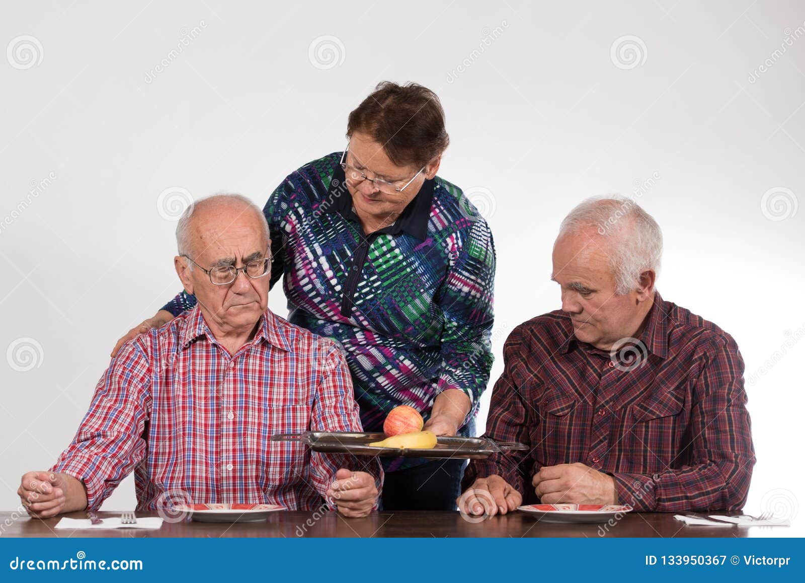 Two men and woman ready for dinner. Two elderly men having discussion, women brings them apple and banana
