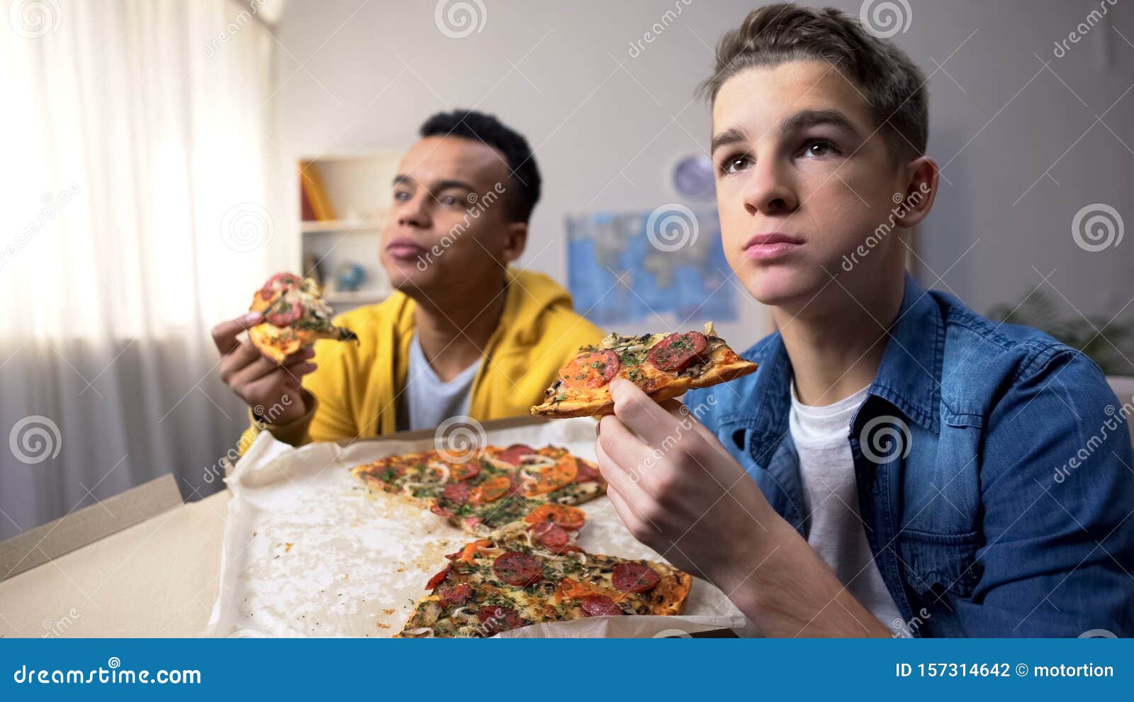 Two Multiethnic Male Teenagers Eating Pizza And Watching Tv Show