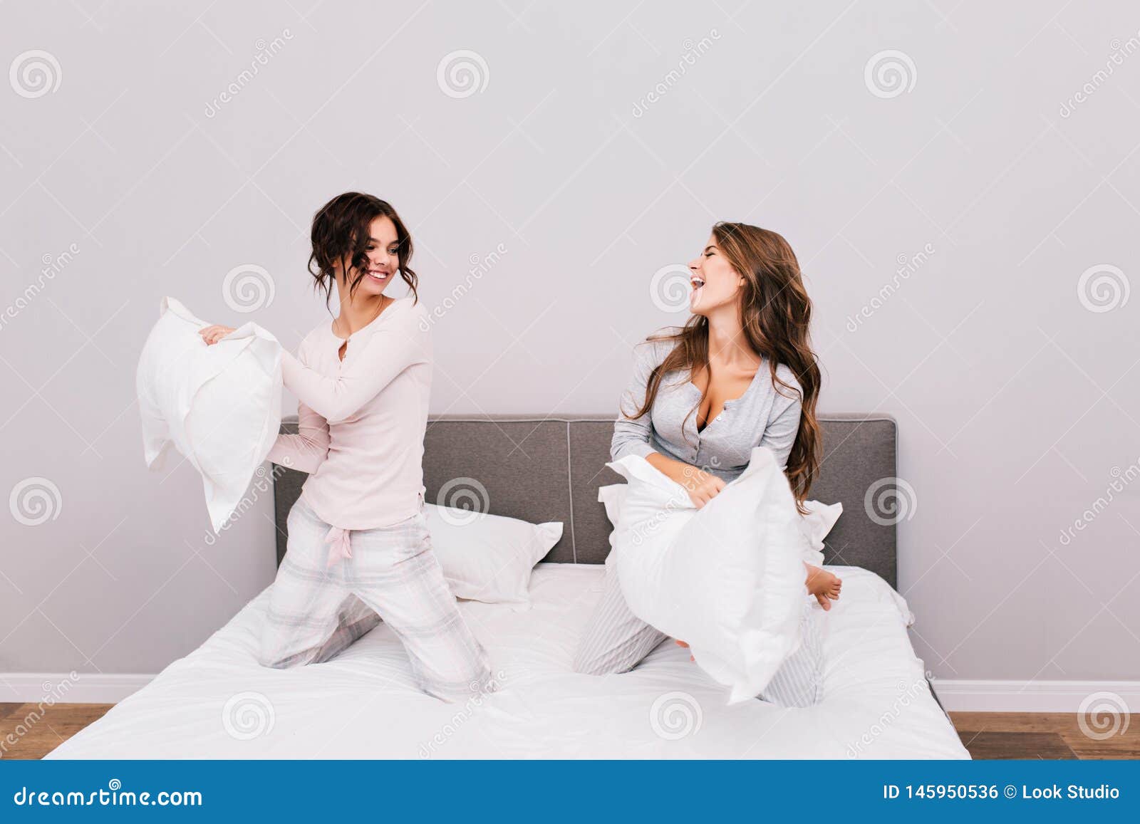 Two Pretty Girls In Pajamas Having Pillow Fight On Bed Stock Photo